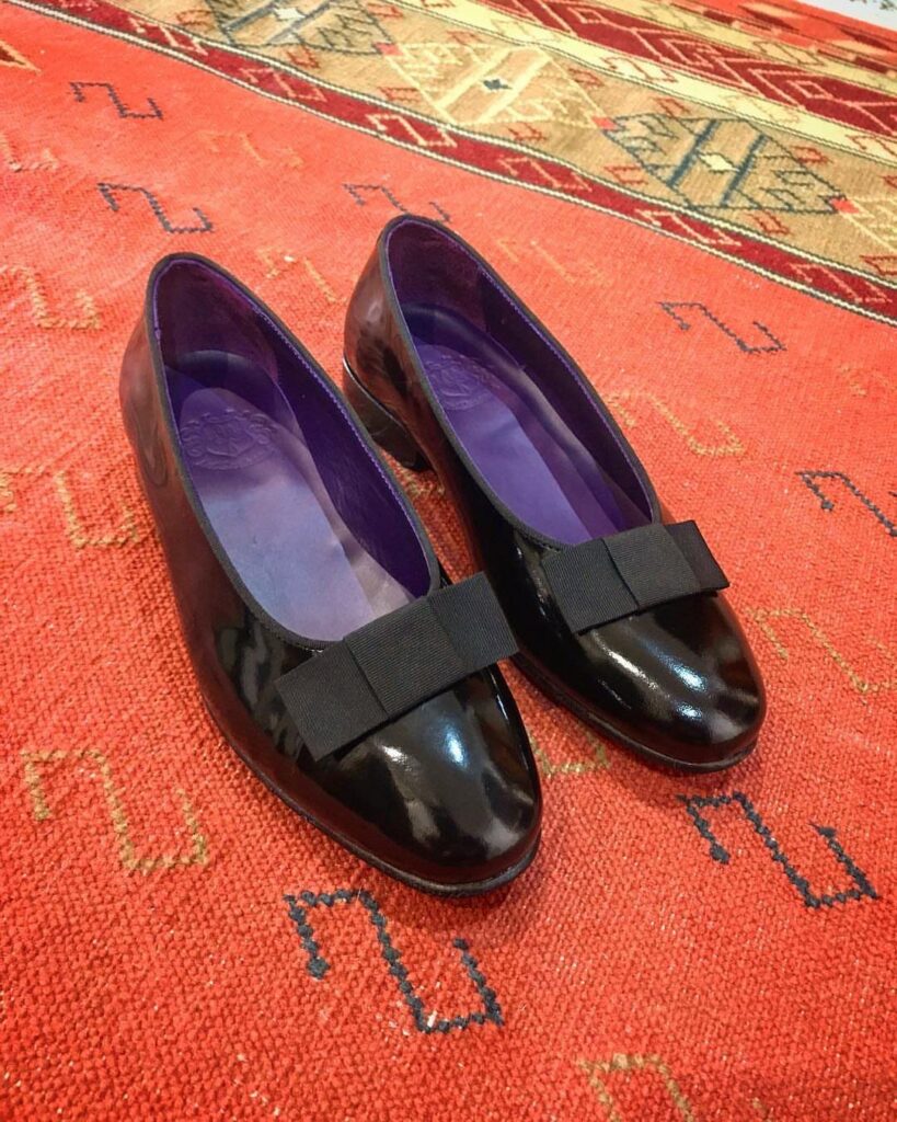 Black patent leather pumps with bows resting on a red patterned rug.
