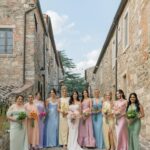 Bridesmaids in various pastel colored formal dresses standing in a stone alleyway holding bouquets.