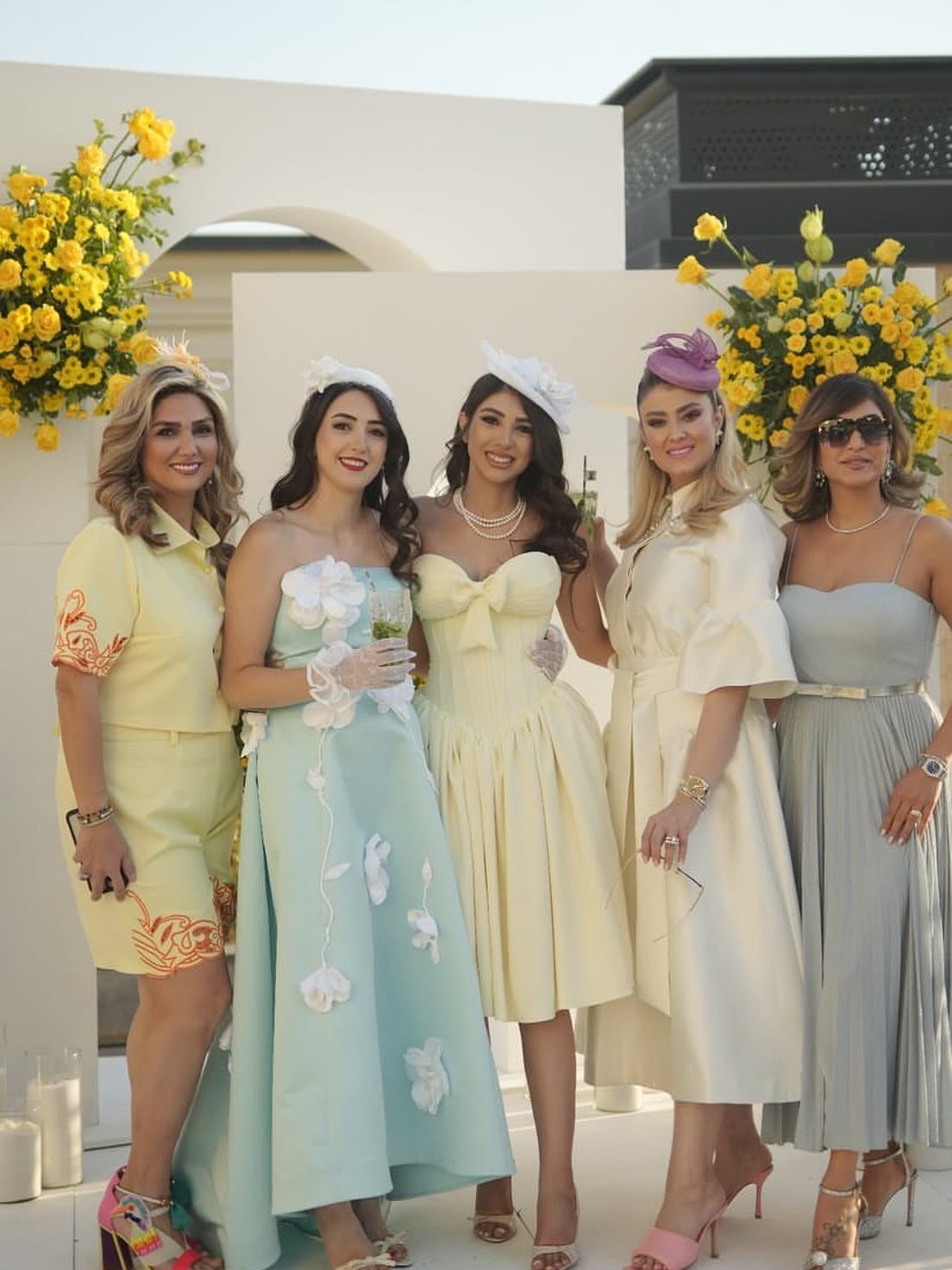 Five women in elegant pastel formal dresses posing together outdoors near yellow flowers.