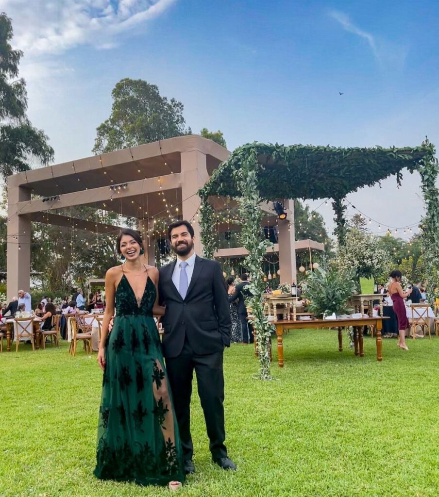 A woman in a dark green lace maxi dress and a man in a suit standing on grass.