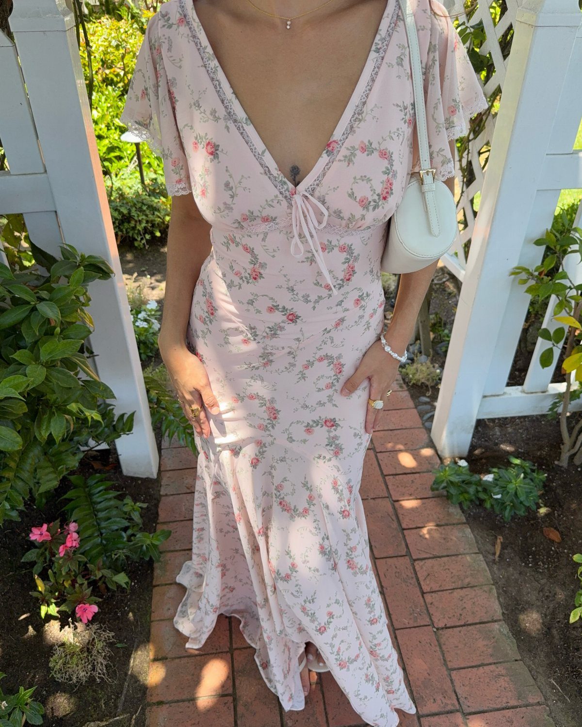 A woman in a pink floral maxi dress standing on a brick path in a garden.