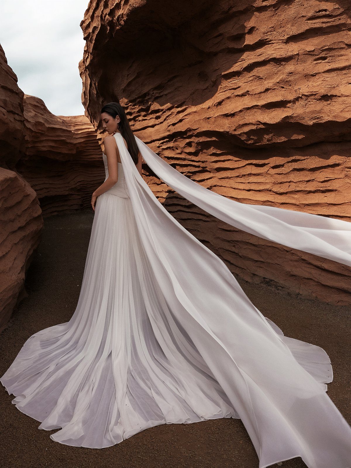 A woman wearing a white pleated wedding dress with a long flowing cape against red rocks.