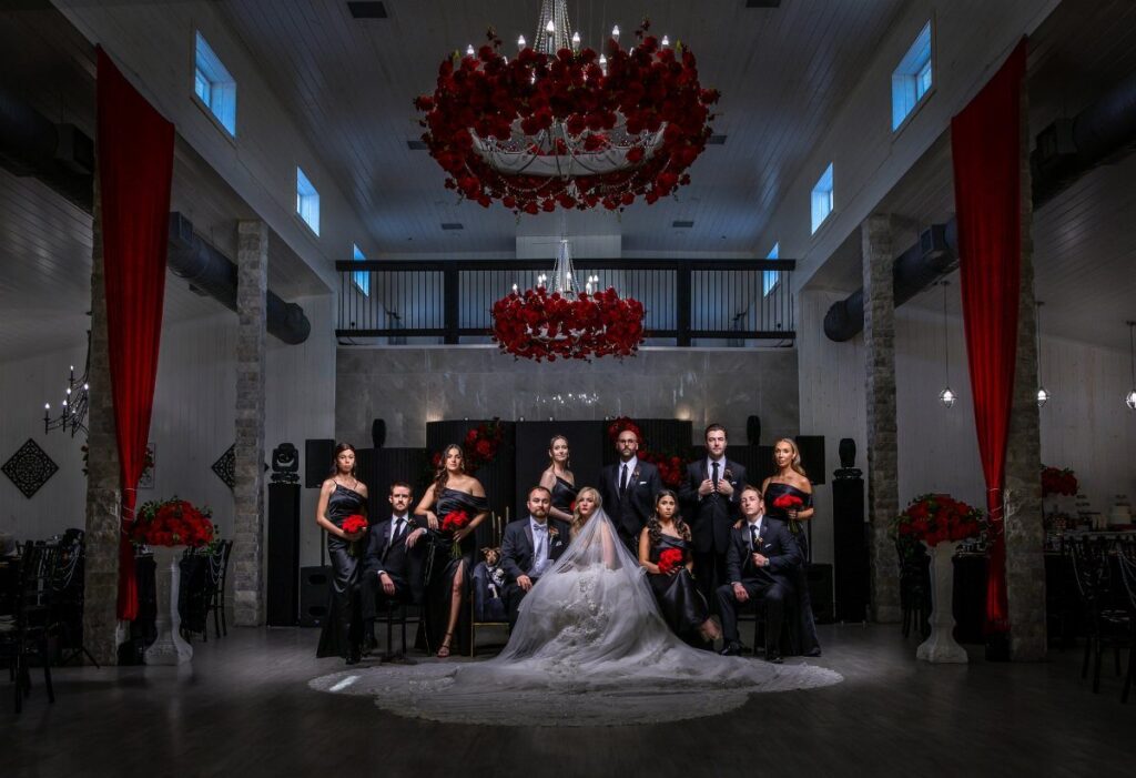 Wedding party in black and red attire posing in a decorated hall with red floral chandeliers.