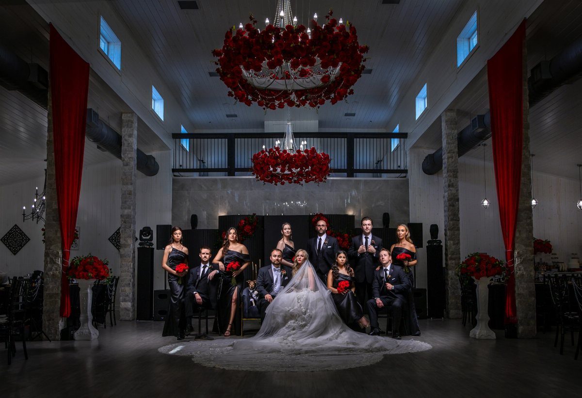 Wedding party in black and red attire posing in a decorated hall with red floral chandeliers.