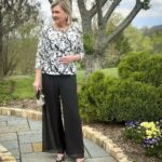 Mature woman in floral print blouse and wide-leg black trousers standing in a garden