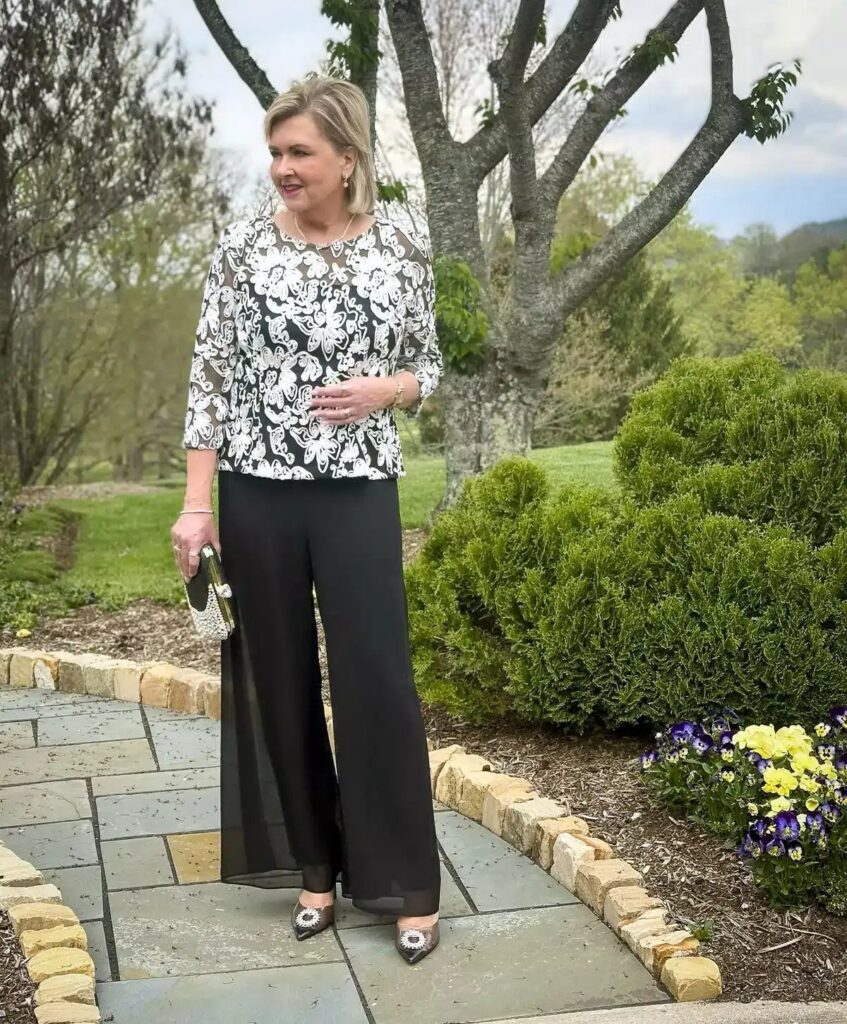 Mature woman in floral print blouse and wide-leg black trousers standing in a garden