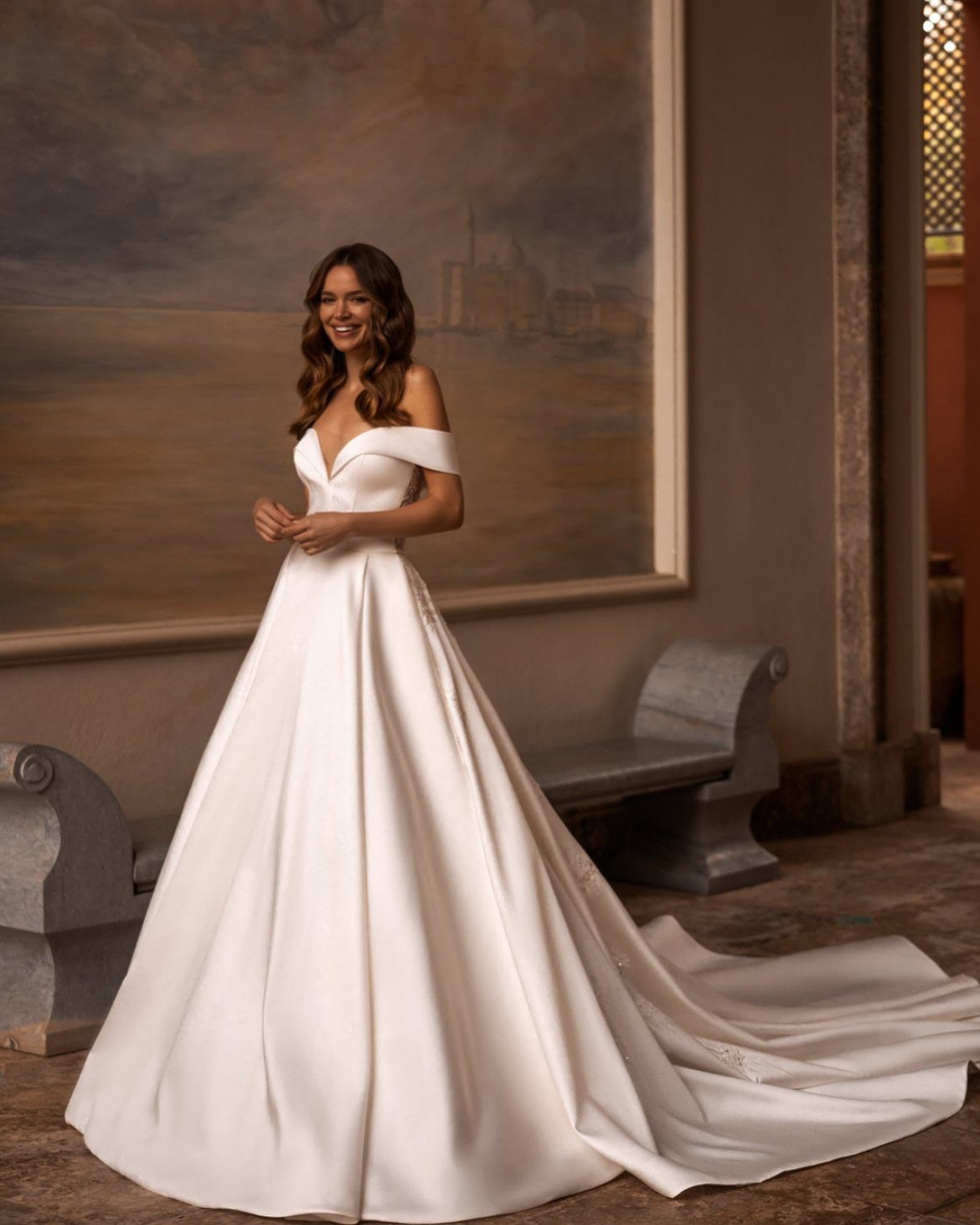 A smiling woman wearing an elegant white off-the-shoulder satin wedding dress with a long train.
