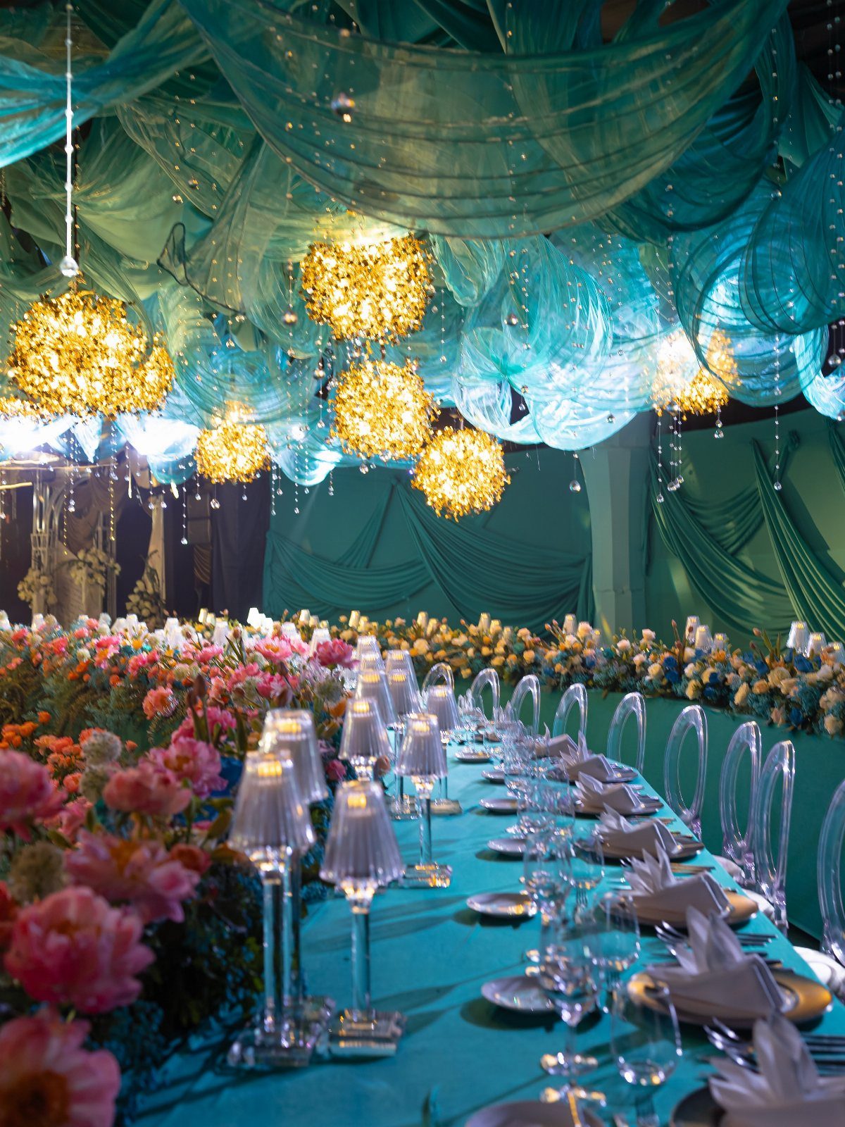 A long dining table with teal linens, pink flowers, and glowing golden chandeliers under draped teal fabric.