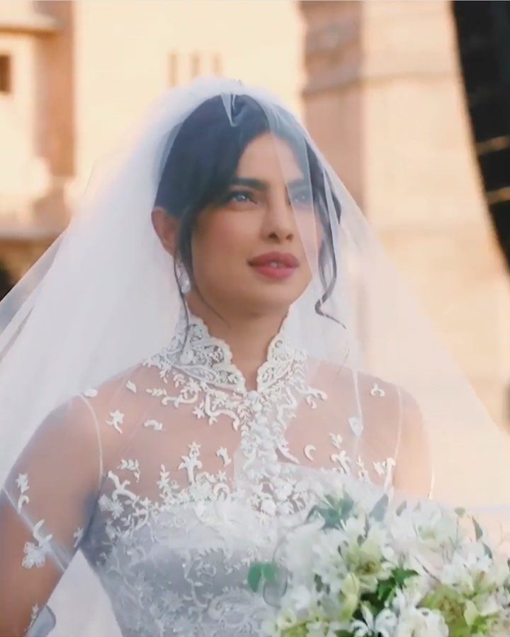 A woman in a white lace wedding dress and sheer veil holding a white flower bouquet.