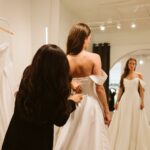 Woman in white wedding dress being fitted by an assistant in a bridal shop.