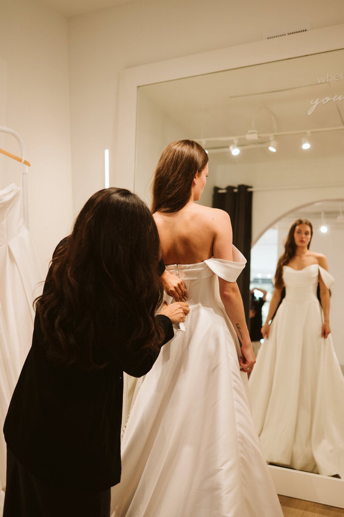 Woman in white wedding dress being fitted by an assistant in a bridal shop.