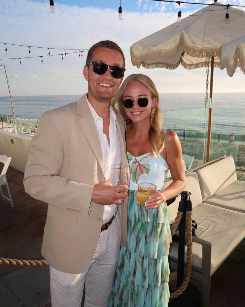 A smiling couple holding drinks stands on a seaside patio overlooking the ocean at sunset.