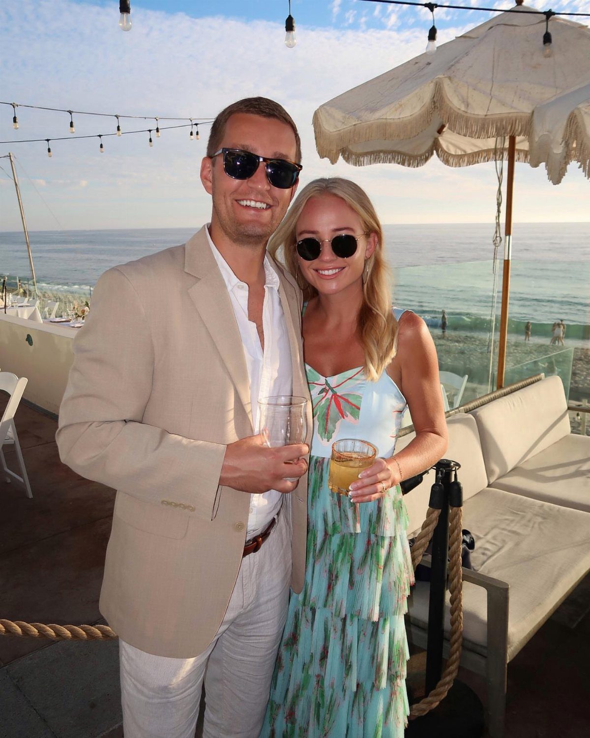 A smiling couple holding drinks stands on a seaside patio overlooking the ocean at sunset.
