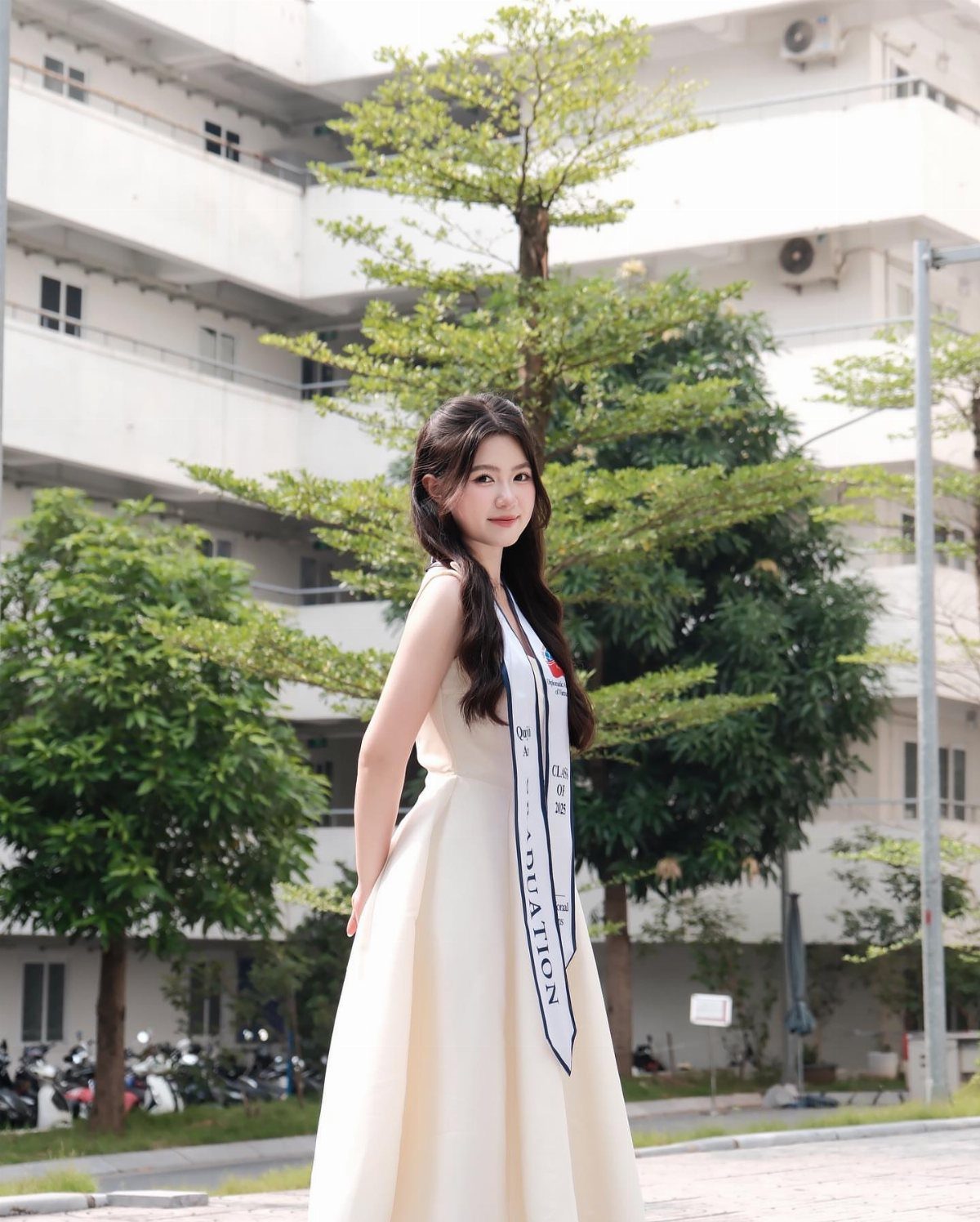 A young woman in a white dress and graduation sash posing outdoors near trees and a building.